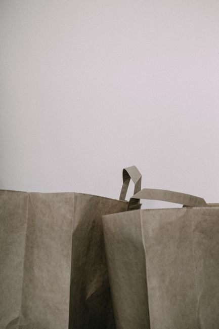 The image shows two large, brown paper shopping bags with twisted paper handles placed against a plain, light-colored wall. The bags are made of kraft paper with a slightly textured surface, and their sides are creased and folded, indicating they are empty or lightly filled. The handles, also crafted from twisted paper, are visible on the bag on the right. The environment appears to be an indoor space with minimal lighting, and the focus of the image is on the paper bags, which are positioned in the lower portion of the frame. These bags could be used for carrying waste or recyclable materials as part of a private rubbish removal service, such as that offered by Rubbish Removal Marylebone, highlighting the importance of proper waste handling and on-site clearance for shops or commercial premises.