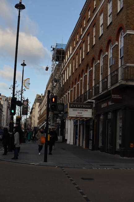 A city street scene in Marylebone showing a row of multi-storey brick buildings with large arched windows and small balconies, some with black railings. The buildings are adjacent to a pavement where pedestrians are walking, including individuals with backpacks and coats. To the left, there are street lamps and traffic lights, with one showing a red pedestrian signal. In the background, some construction scaffolding is visible on one of the buildings, indicating ongoing work. The street appears to be a busy commercial area, and the lower level of the buildings includes storefronts, one of which displays a sign for Marylebone. The natural daylight illuminates the scene, suggesting late afternoon, with a partly cloudy sky overhead. This urban setting reflects a typical Marylebone shopping street, where independent collection and private disposal of waste may be relevant, aligning with services like rubbish removal from businesses and shops in the area, as provided by Rubbish Removal Marylebone.