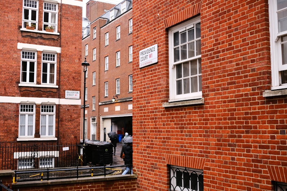 A narrow urban alleyway between red-brick residential buildings in Marylebone, with white-framed sash windows and decorative brickwork. Two individuals are seen at the rear near a large black wheelie bin and a smaller container, possibly engaged in rubbish collection or disposal activities. The scene includes a traditional street lamp on a black metal post, and street signs indicating 'Balderton Street W1' and 'Providence Court W1,' mounted on the facades of the buildings. The environment appears tidy, with clean brickwork and minimal clutter, suggesting regular waste management. The lighting is soft and natural, indicating daytime conditions, and the overall setting aligns with independent waste removal services provided by companies like Rubbish Removal Marylebone, supporting local rubbish clearance or private rubbish collection practices in the area.