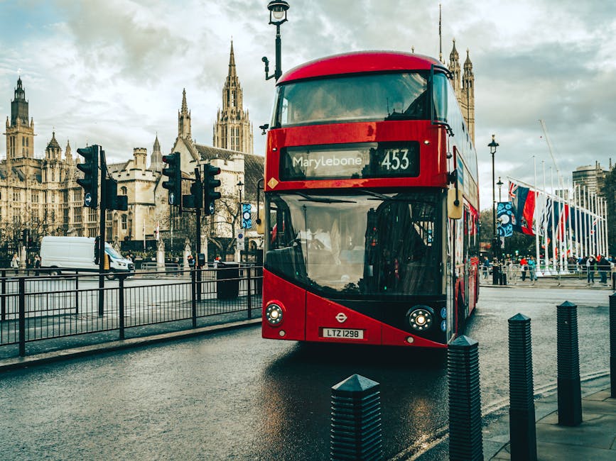 A red double-decker bus with the route number 453 and destination Marylebone is approaching along a wet city street in front of Westminster Abbey, visible in the background with its Gothic spires and detailed stone architecture. The bus has a modern, curved front with a large windshield, and features the London Transport logo on the lower front panel, along with a white registration plate reading LTZ 1298. To the left of the bus, there are a series of black pole-mounted bollards separating the roadway from the sidewalk. Several black traffic lights are positioned along the street, and in the background on the right, there are Union Jack flags hanging on poles near a pedestrian area filled with a few people walking or waiting. The scene appears to be late afternoon or early evening, with cloudy skies reflecting off the wet asphalt, creating a slightly subdued atmosphere, relevant to urban rubbish collection and city logistics, typical of independent waste removal services like Rubbish Removal Marylebone.