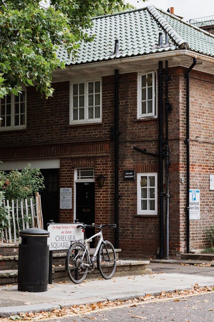 A two-storey brick building with a light grey tiled roof and white-framed sash windows on both floors. In front of the building, on the pavement, there is a black cylindrical waste bin positioned next to a white bicycle with a black seat and handlebars, leaning against a small white sign attached to a low metal fence. The sign indicates the location as Chelsea Square, and there is a small informational noticeboard mounted on the building’s wall beside the front door. The surrounding area is shaded by a leafy green tree on the left, and autumn leaves are scattered across the sidewalk. To the right side of the building, a white parking sign and a blue disc parking zone sign are attached to the wall. Black drainpipes run vertically along the façade, and the environment suggests an urban residential area with nearby greenery, consistent with private waste handling or alternate rubbish collection services that may be managed independently of local authority schemes.