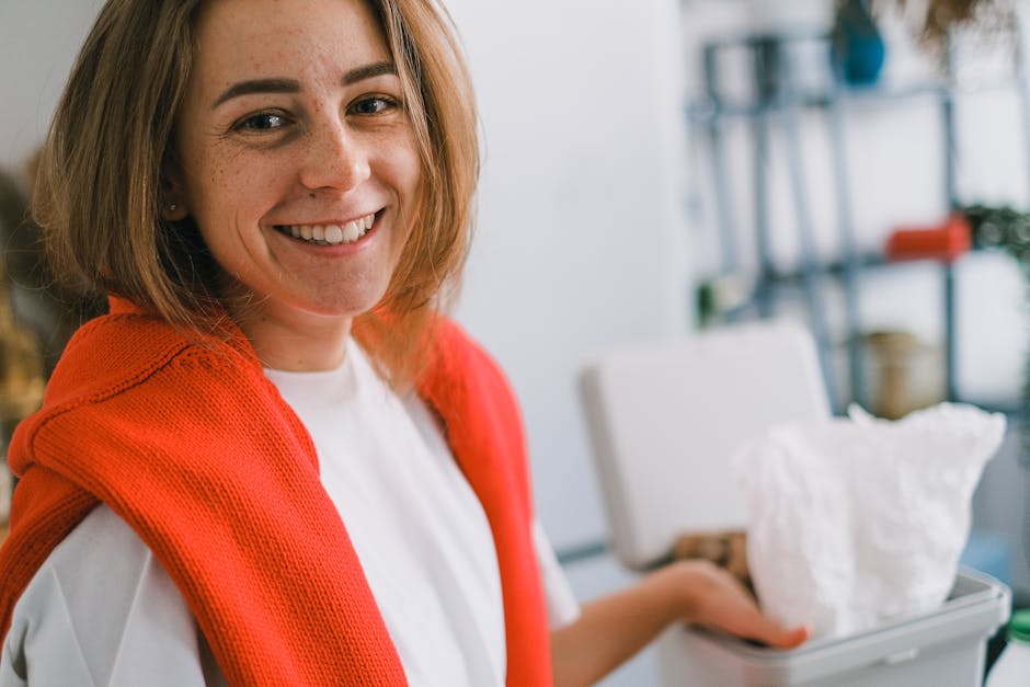 A woman with shoulder-length light brown hair, fair skin with freckles, and a cheerful expression is smiling while standing indoors. She is wearing a white shirt and has a red knitted cardigan draped over her shoulders. In the background, a white plastic bin or container holds crumpled white paper or tissue, which she is lifting or placing inside. Behind her, a metal shelving unit is visible with several shelves holding various items such as blue and green containers, a plant, and other miscellaneous objects. The environment appears well-lit with natural or soft indoor lighting, and the scene suggests an informal setting related to waste management or recycling, aligning with activities like rubbish disposal or on-site waste handling, as part of services offered by Rubbish Removal Marylebone.