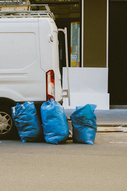 A white commercial van parked on a street during daylight hours, with its rear door partially open and three large blue rubbish bags placed on the pavement beside it. The bags are made of a plastic material with a slightly crinkled texture and are filled with waste, leaning slightly to one side. The background shows the exterior of a building with a glass window and a white panel section, suggesting an urban environment suitable for private waste collection or rubbish removal services. The scene depicts a typical setup for independent rubbish disposal, with the bags ready for collection by a waste removal company like Rubbish Removal Marylebone, emphasizing the process of on-site clearance outside a commercial or residential property.