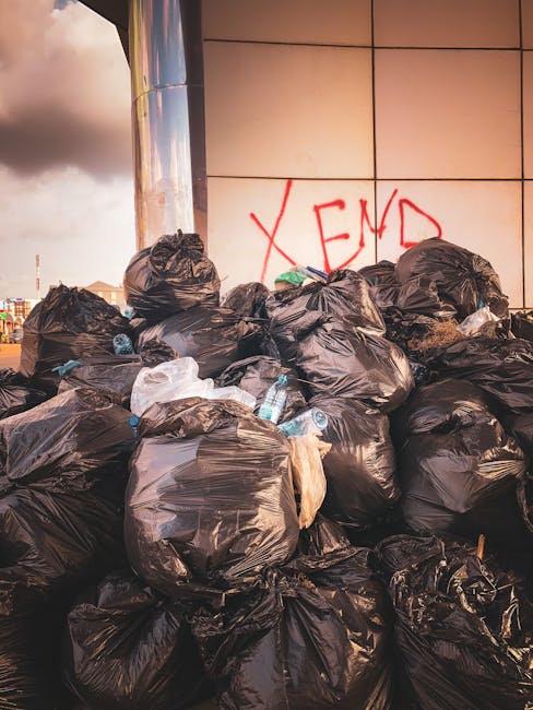 A large pile of black plastic rubbish bags filled with waste is stacked against a modern building with a tiled exterior wall. Some bags are torn, revealing glimpses of plastic bottles and other refuse inside. The waste is situated on a paved surface, possibly on a street or urban area, with the bags crowded together and extending to the lower part of the image. In the background, the tiled wall has graffiti with the word 'XEND' spray-painted in red. The lighting suggests late afternoon or early evening, with warm tones casting a slight glow over the scene. This image highlights private waste accumulation outside a commercial or residential building, illustrating the importance of alternative waste handling or independent removal services to prevent excess rubbish build-up, as might be offered by rubbish removal companies like Rubbish Removal Marylebone.
