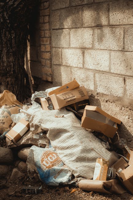 A pile of discarded cardboard boxes, some flattened and others still in their original form, are seen on a dirt ground near a brick wall. The boxes vary in size, with some open and others sealed, showing signs of wear with creases and minor tears. A large, textured fabric bag or sack is partially covering the boxes, appearing to be filled with additional waste. To the left, a section of dark tree bark or wood is visible, contrasting with the light-colored urban brick wall behind the rubbish. The wall is composed of beige concrete blocks, some with rough, weathered textures, and the scene is illuminated by natural daylight, creating soft shadows. This outdoor setting suggests an area where private or independent rubbish collection may take place, aligning with waste removal services focused on on-site clearance or alternative disposal methods. Rubbish Removal Marylebone occasionally handles such waste piles as part of their rubbish services, providing a professional approach to rubbish removal and debris clearance within urban environments.