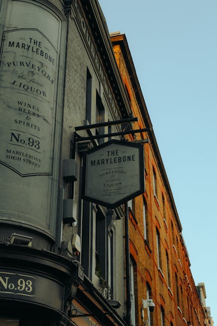Photograph of a traditional pub sign hanging from the exterior of a brick building, with the sign reading 'The Marylebone' and advertising wine, spirits, and liquors. The sign is black with white lettering and is attached to a black metal bracket extending from the building's façade. The building has a warm, orange-brown brick finish with multiple windows and a slightly weathered appearance, typical of historic urban architecture. Behind the sign, the light blue sky indicates daytime, and the scene suggests a street in a busy area. The setting reflects an environment where local businesses, such as pubs and shops, contribute to the community ambiance, often requiring occasional rubbish removal services to maintain cleanliness in commercial districts. Rubbish Removal Marylebone operates in such areas to support the ongoing management of waste from these retail and hospitality establishments, ensuring tidy streets and proper disposal of waste materials.