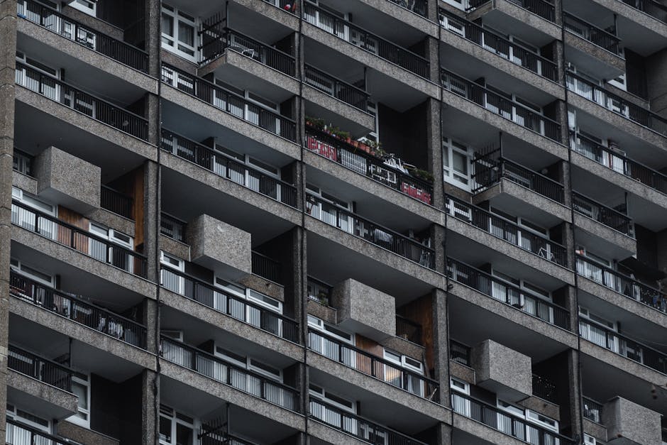 A close-up view of a multi-storey residential building's exterior façade, showing numerous balconies with black metal railings and concrete surfaces. The balconies are arranged in a grid pattern, with some featuring potted plants, outdoor furniture, or laundry hanging. The building's façade is constructed with rough textured grey concrete, with some sections displaying a mixture of darker and lighter tones. Several windows with white frames and glass panes are visible between the balconies, reflecting ambient light. The overall scene is illuminated by natural daylight, highlighting the geometric repetition of the balconies and the utilitarian design typical of urban apartment blocks. This image conveys an environment where tenants regularly manage their outdoor spaces, aligning with the context of on-site waste management or private rubbish collection outside high-rise flats, as handled by companies like Rubbish Removal Marylebone.
