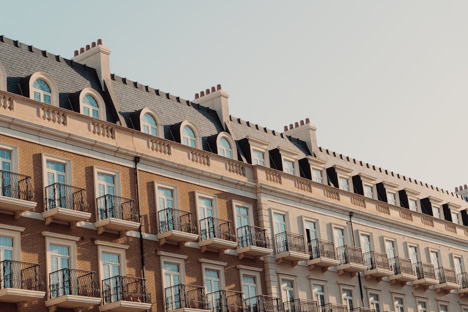 A close-up view of the upper floors of a multi-storey residential building with a classic architectural style, featuring a light beige and grey colour scheme. The building has rows of uniformly spaced windows with white frames, some of which open onto small decorative balconies with black metal railings. The roof is dark grey with dormer windows and chimney stacks evenly spaced along the ridge. The exterior walls display a combination of brickwork and ornamental mouldings, and the overall scene is illuminated by natural daylight. This setting suggests an urban environment suitable for private waste management or independent rubbish collection services, like those offered by Rubbish Removal Marylebone, in line with the context of reducing clearance disruption for flats on W1U estates.