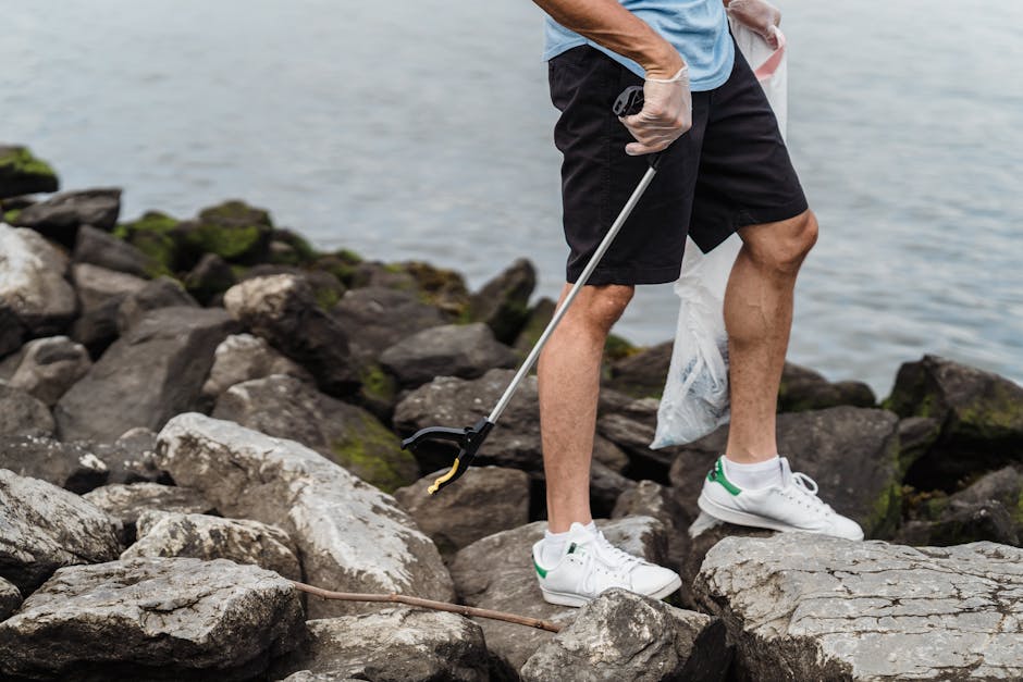 The image shows a person standing on a rocky shoreline near a body of water, with their lower body visible from the waist down. They are dressed in black shorts, a light blue t-shirt, and white sneakers with green accents. The individual is wearing gloves and holding a litter picker, using it to collect trash among the large, uneven rocks. The rocks vary in size and color, predominantly shades of grey and brown, with some areas covered in patches of green moss or algae. The background features calm water, indicating a natural outdoor environment. The scene suggests a clean-up activity, such as an environmental or rubbish removal effort, with the person actively engaged in clearing debris from the shoreline. This visual context aligns with private or community-based waste management services that focus on on-site rubbish collection and environmental preservation.