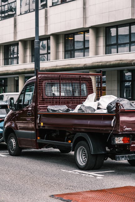 A small burgundy utility truck is parked on an urban street in front of a modern multi-storey building with large glass windows and a concrete facade. The truck's cargo bed is filled with a variety of waste items, including several large bags made of white and grey plastic, as well as some loose, indistinct debris. The vehicle has a protective metal grid behind the cab, likely to secure the load, and visible rear lights and black tires with silver rims. Surrounding the truck, other parked vehicles are visible along the street, and a tall streetlamp stands to the left of the image. The scene appears to be part of a private waste collection or on-site clearance operation, related to the services offered by Rubbish Removal Marylebone, which may involve alternative waste handling approaches outside of standard council rubbish collection. The lighting is natural, indicating daytime, and the overall environment is typical of a city centre setting focused on waste removal or rubbish disposal activities.