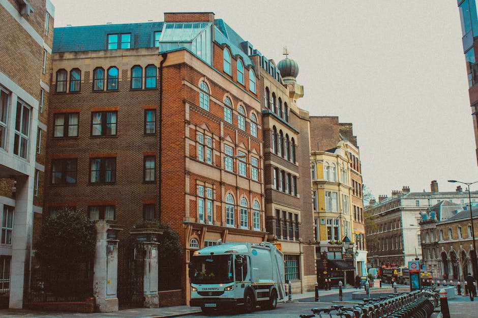 A white rubbish collection truck with a blue logo on its side is positioned on a city street with brick and stone buildings in the background. The truck has a closed rear compartment and is parked adjacent to a sidewalk paved with concrete slabs. The surrounding buildings display a mix of architectural styles, including red brick, beige stone, and ornate facades, with some featuring arched and sash windows. The street appears to be in an urban area with a few bollards, bicycle racks, and a street lamp visible along the curb. The scene is captured during daylight with soft, overcast lighting, and the overall environment suggests an active city center where local waste management services such as Rubbish Removal Marylebone may operate for private rubbish collection and on-site clearance of waste materials.