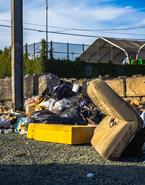 A close-up view of a roadside pile of discarded rubbish including black trash bags, a yellow plastic container, a worn-out beige car seat with visible fabric texture, and miscellaneous small waste items. The debris is situated on a gravel surface adjacent to a stone wall made of irregularly shaped stones. In the background, there is a metal fence, some green foliage, and a partly cloudy blue sky. Overhead power lines stretch across the scene, and a large, curved shelter structure with a white roof is visible behind the fence. The overall scene illustrates a typical example of illegally dumped waste that may require professional rubbish removal services, such as those provided by Rubbish Removal Marylebone, to facilitate proper clearance and disposal.
