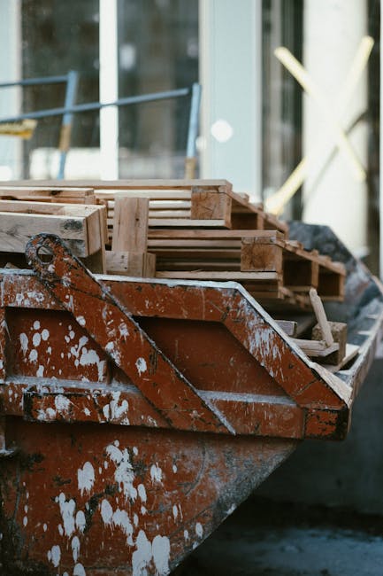 A close-up view of an old, rusted orange metal boat with visible white paint splatters, docked outdoors next to a building with large glass windows. Resting inside the boat are several wooden pallets and planks, which have a rough texture and natural wood finish, with some showing signs of weathering and minor splintering. The pallets are stacked unevenly, with the top ones partially leaning to one side. The background shows a partially visible glass-fronted building, suggesting an urban or commercial setting, with some metal framework and blurred foliage visible through the glass. The overall scene is lit by natural daylight, highlighting the weathered surface of the boat and the textures of the wooden materials, subtly illustrating a scene related to site clearance or material staging, common in waste removal or rubbish collection contexts.