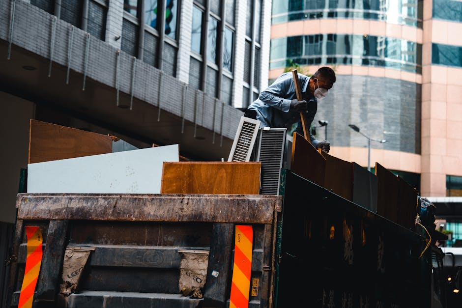 A man wearing a grey shirt, dark trousers, black gloves, and a face mask is standing on the edge of a large, open-top waste truck, positioned in an urban environment with tall modern office buildings in the background. He is leaning forward slightly, lifting or adjusting a piece of discarded furniture or large object among other debris. The truck bed contains various items including wooden panels, a small white radiator, and a piece of white board or panel, all showing signs of wear and grime. The surface of the truck is rusted with visible dirt, and the back of the truck has bright red and yellow reflective safety markings. The scene is lit by natural daylight, with shadows cast on the surrounding buildings, illustrating a typical private waste collection or rubbish removal process in a metropolitan setting associated with independent rubbish disposal services offered by companies like Rubbish Removal Marylebone, supporting alternative waste handling methods outside of standard council rubbish collection. The vertical walls of the truck and the urban setting emphasize the practical nature of waste loading activities, focusing on the collection of bulky refuse in a commercial or residential district.