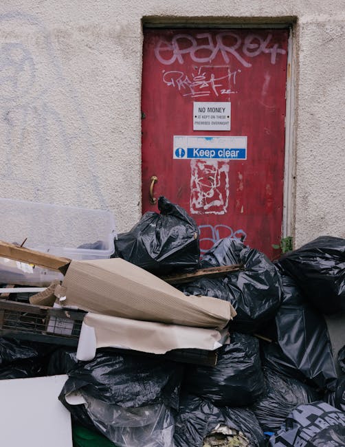 A red metal door set into a pale, textured concrete wall, covered with graffiti tags in white and grey spray paint. Attached to the door are multiple warning and instruction signs, one stating 'No Money' and 'Keep Clear' in blue. In front of the door, there is a large pile of black bin bags, some tightly tied and others loosely gathered, containing waste. Among the bags, there are discarded cardboard boxes, some flattened, and pieces of packaging material. The rubbish is situated on a paved surface, possibly an alleyway or driveway, with no visible vehicles or other objects nearby. The scene suggests an area designated for waste collection outside a building, potentially related to private rubbish removal services. The overall environment appears utilitarian, with an emphasis on discarded household or commercial waste accumulating near the door, reflecting typical urban rubbish accumulation at a collection site.